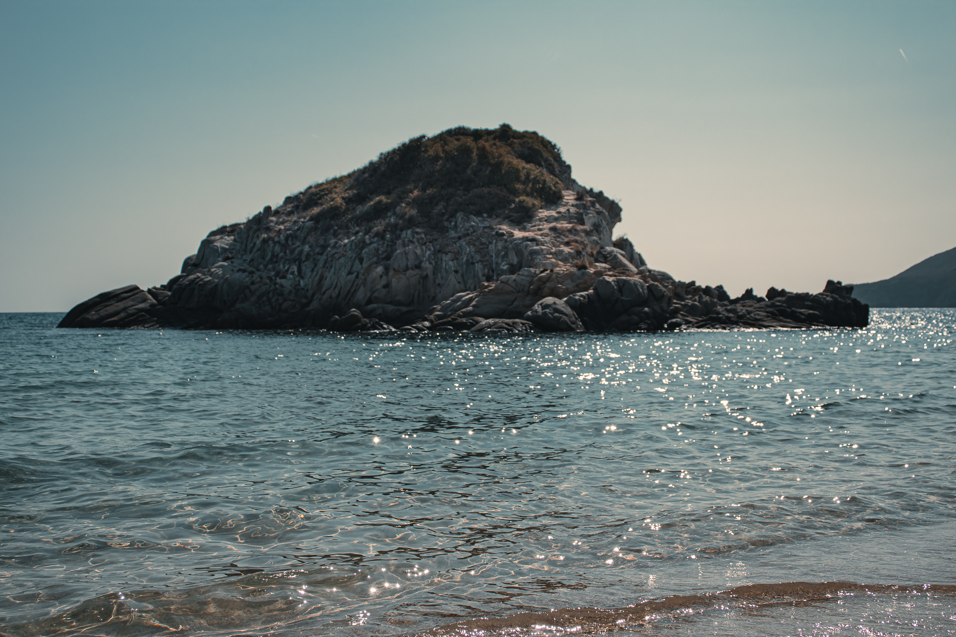 Rocky island rising from calm blue sea with sparse vegetation and sunlit water around it
