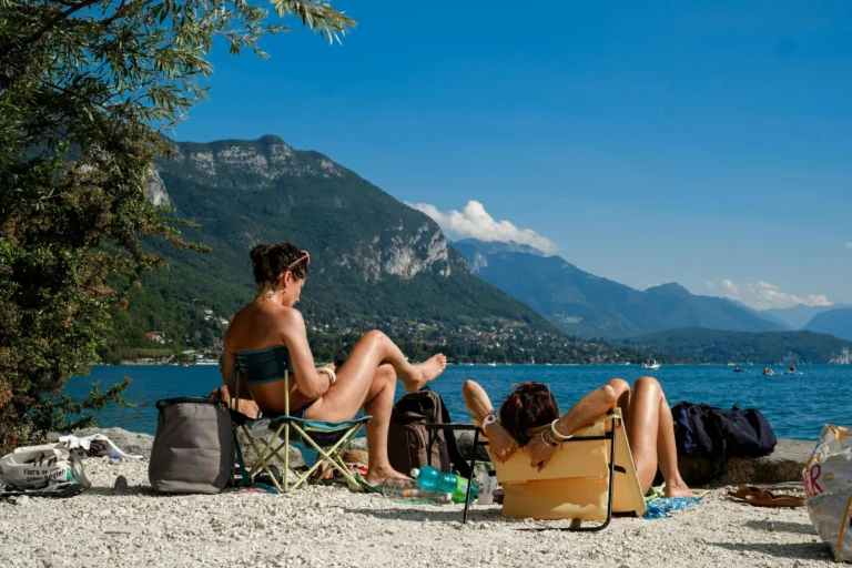 Sunbathers relaxing on a rocky lakeside beach with mountains and blue sky in the background