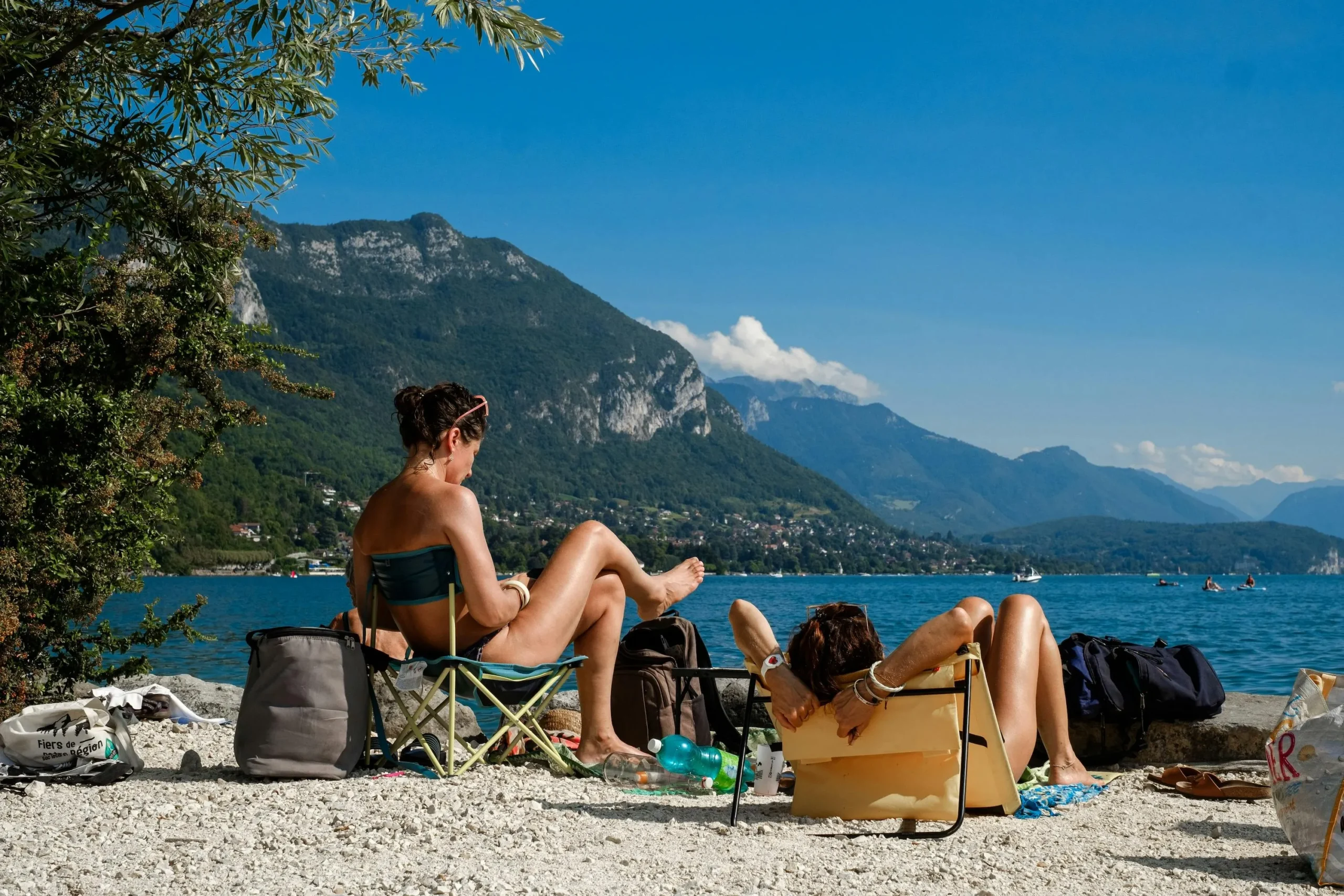 Sunbathers relaxing on a rocky lakeside beach with mountains and blue sky in the background
