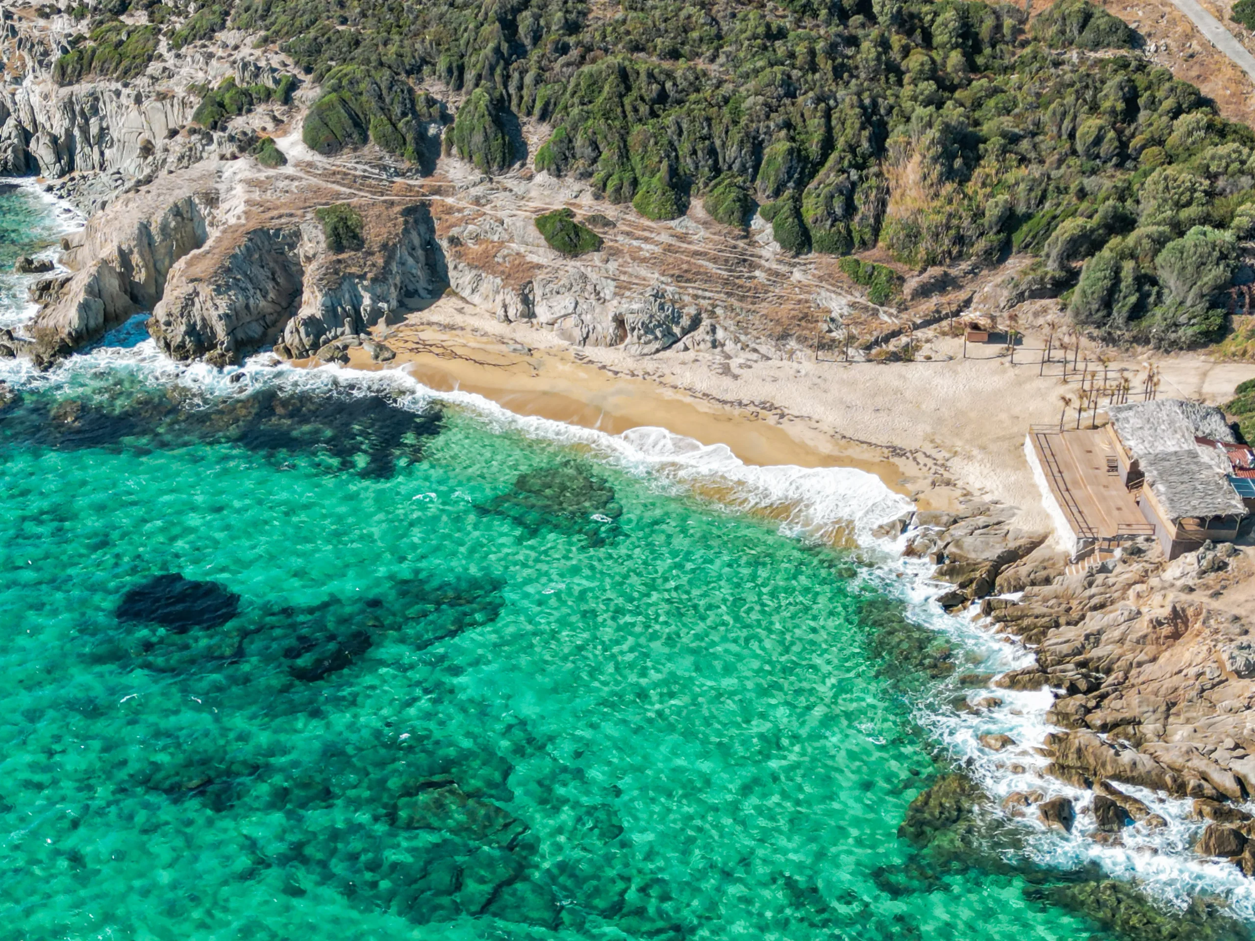 Aerial view of a rocky coastline with bright turquoise water, a sandy beach, and dense green vegetation along the shore.