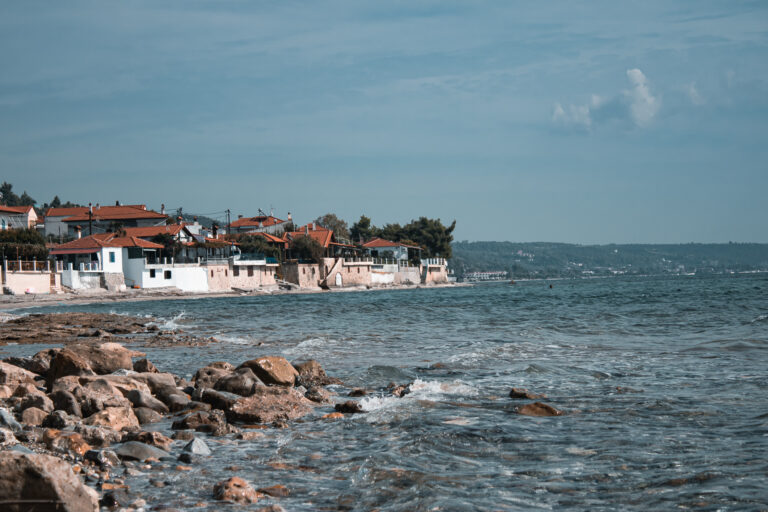 Coastal village with white buildings and red-tiled roofs along a rocky shoreline by the sea under a blue sky.